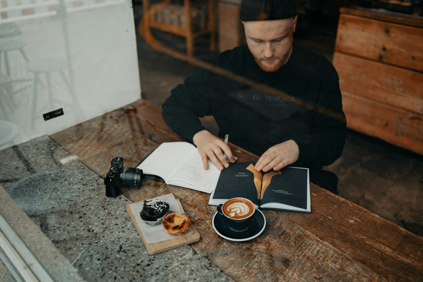 Man writing in notebook at cafe table with pastries and coffee.